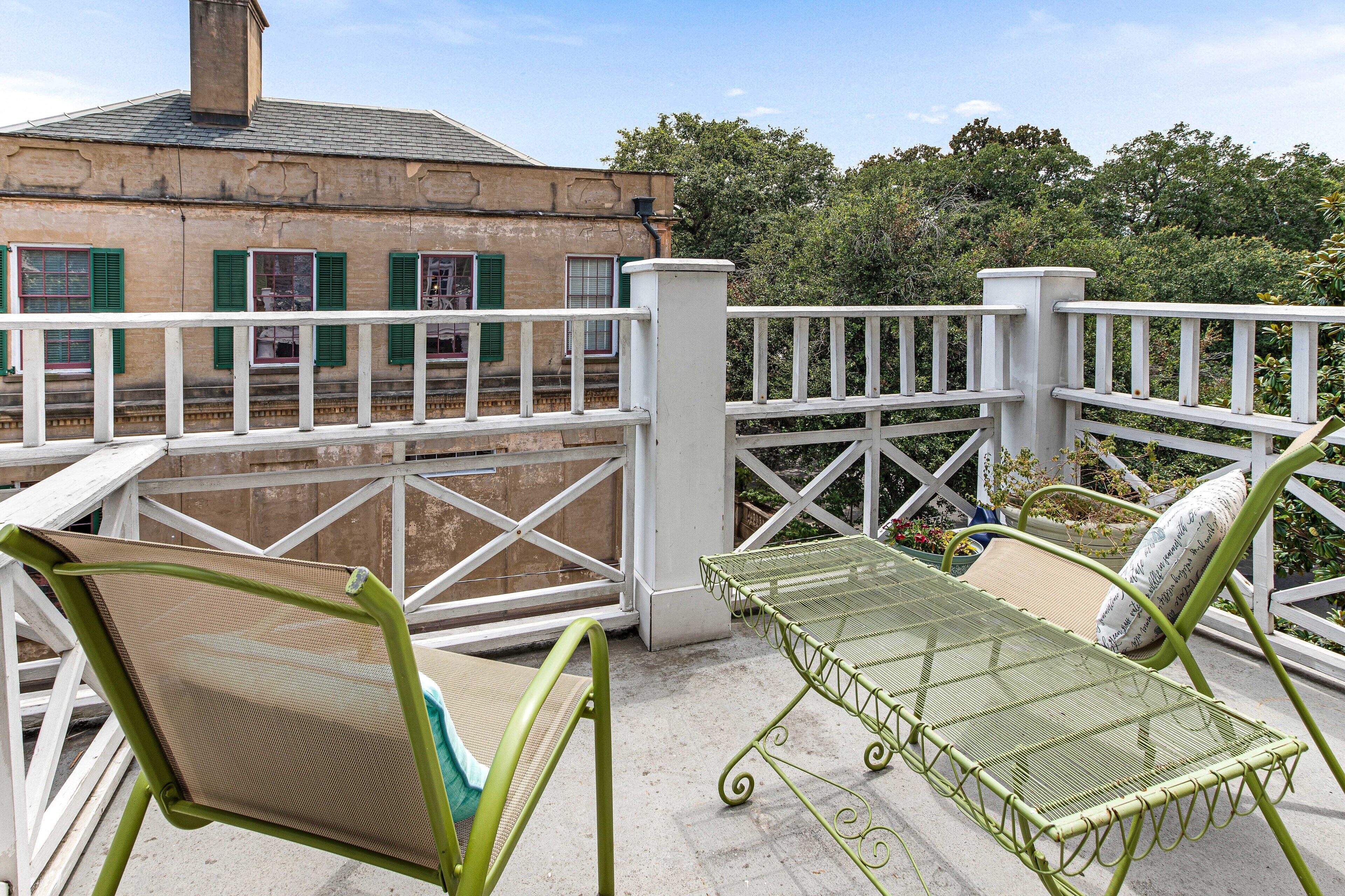 A balcony with a metal table and chairs, a white railing, and a building with green shutters in the background.