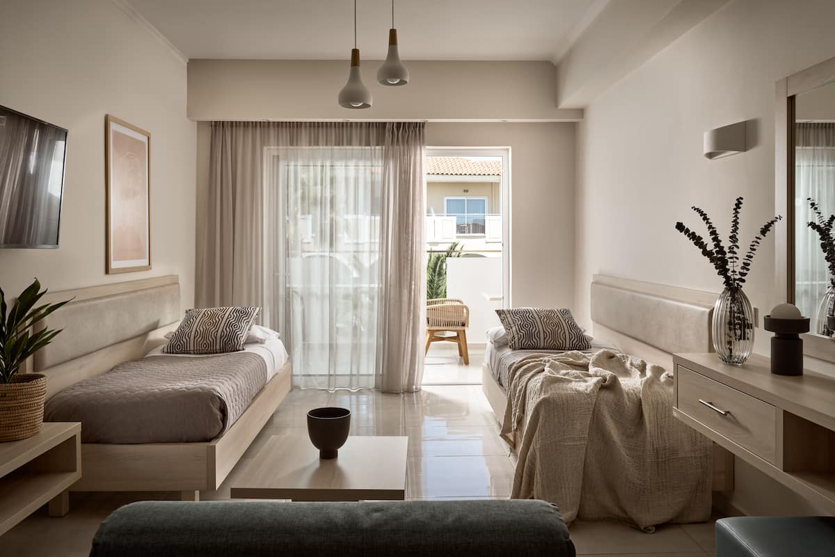 A modern bedroom with a bed, bedside tables, a sofa, and a small table. There is a window with curtains, a plant, and a vase with flowers.