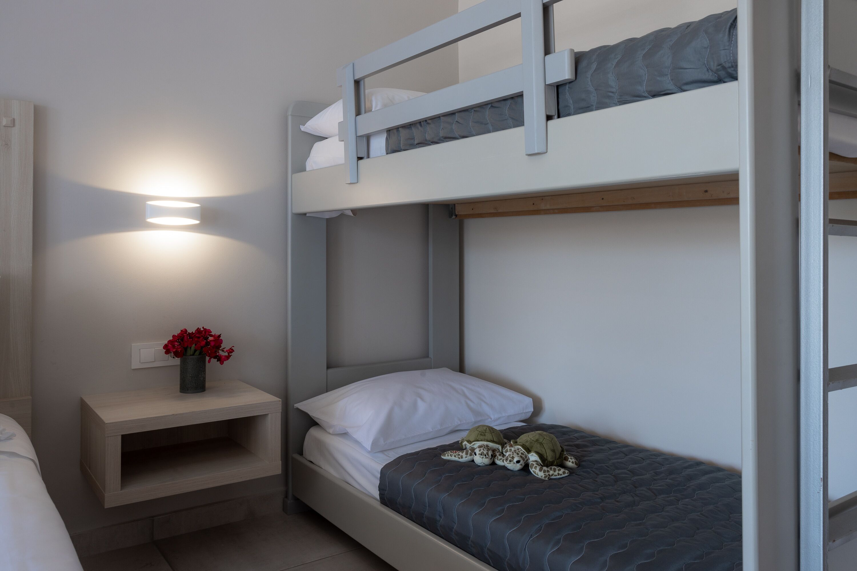 A bunk bed room with a bedside table, a wall-mounted light, and a vase with flowers.