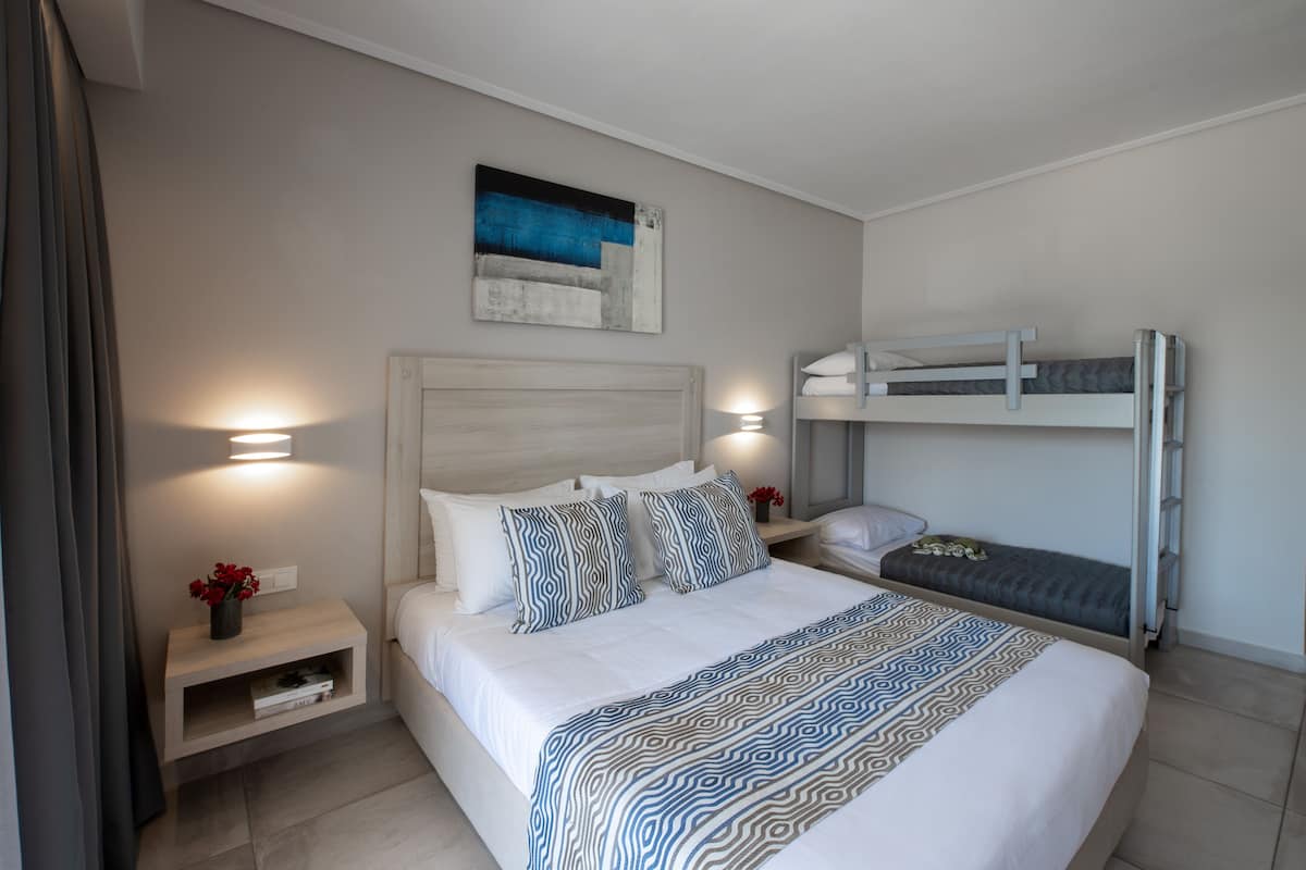 A modern hotel room with a large bed, a bunk bed, a bedside table with a lamp, and a wall-mounted artwork.