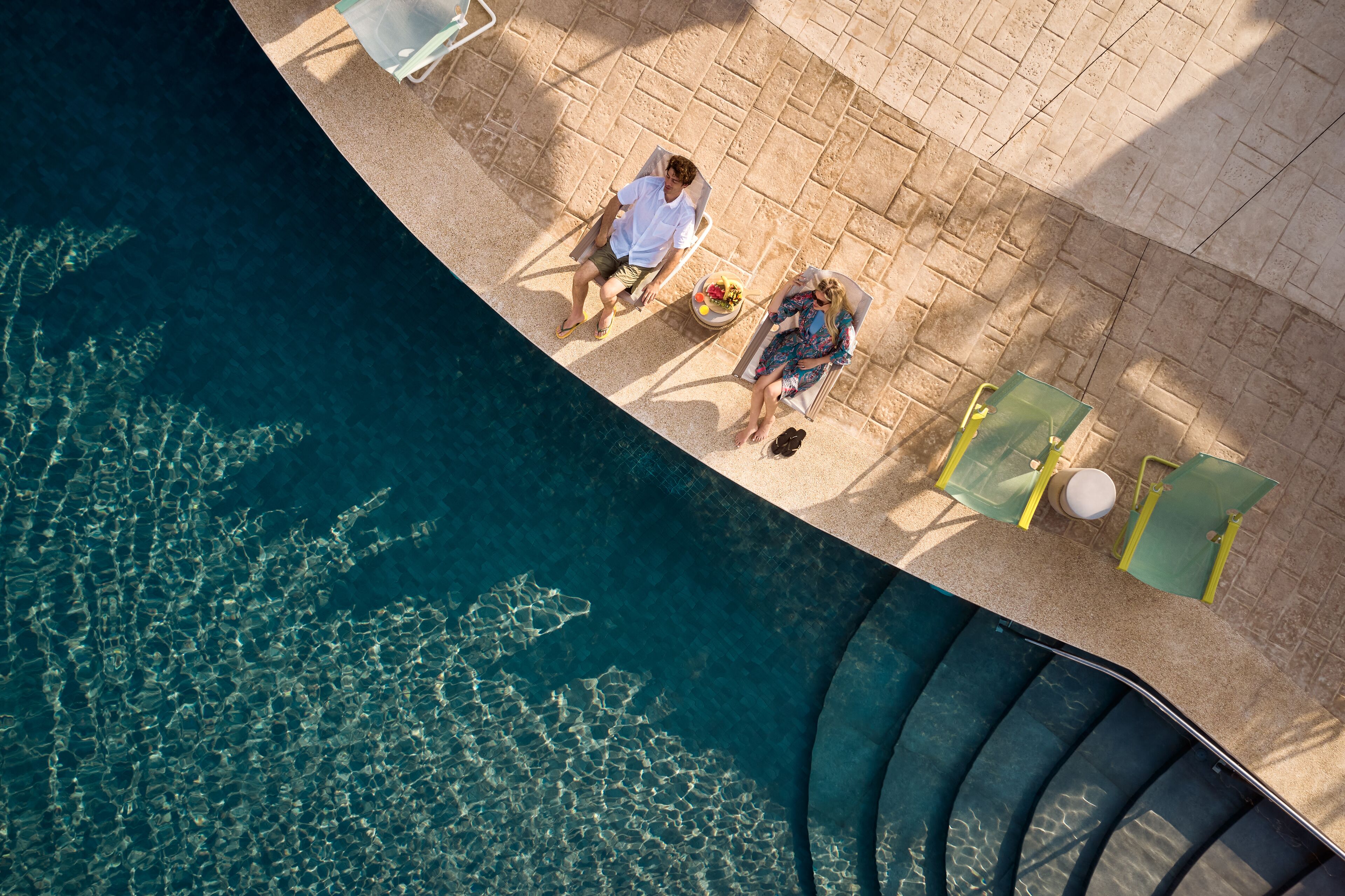 An aerial view of a pool area with two lounge chairs, a tiled deck, and clear water.