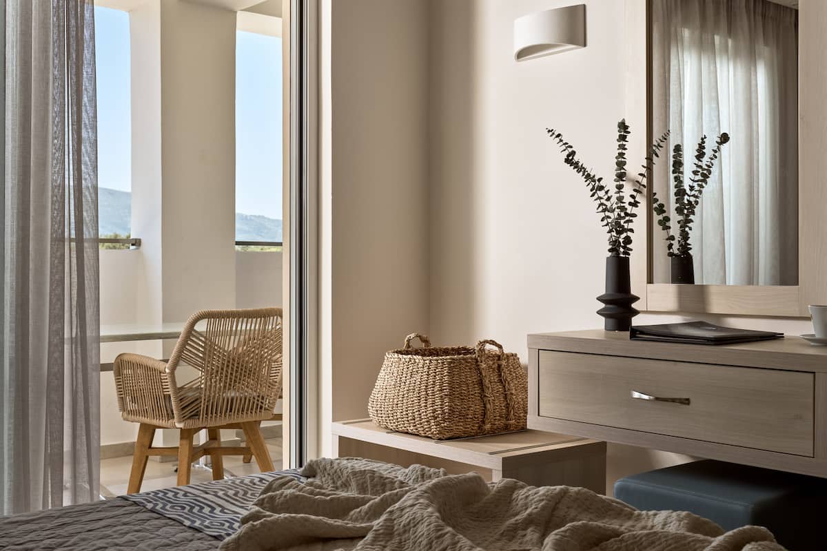 A bedroom with a bed, a bedside table with a lamp, a chair, a wicker basket, and a view of the outdoors.
