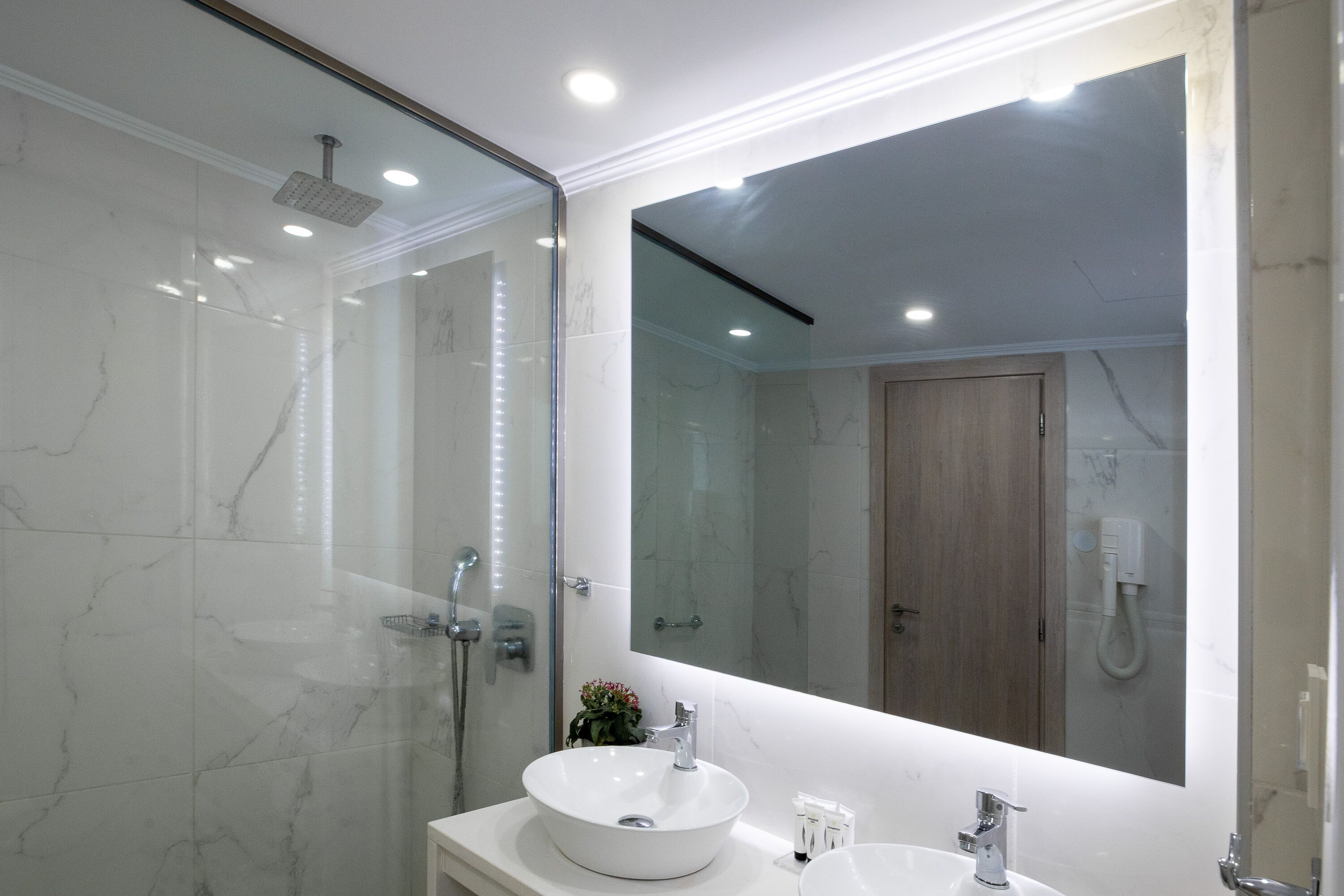 Un baño moderno con un espejo grande, un lavamanos blanco y una zona de ducha con paredes de vidrio.