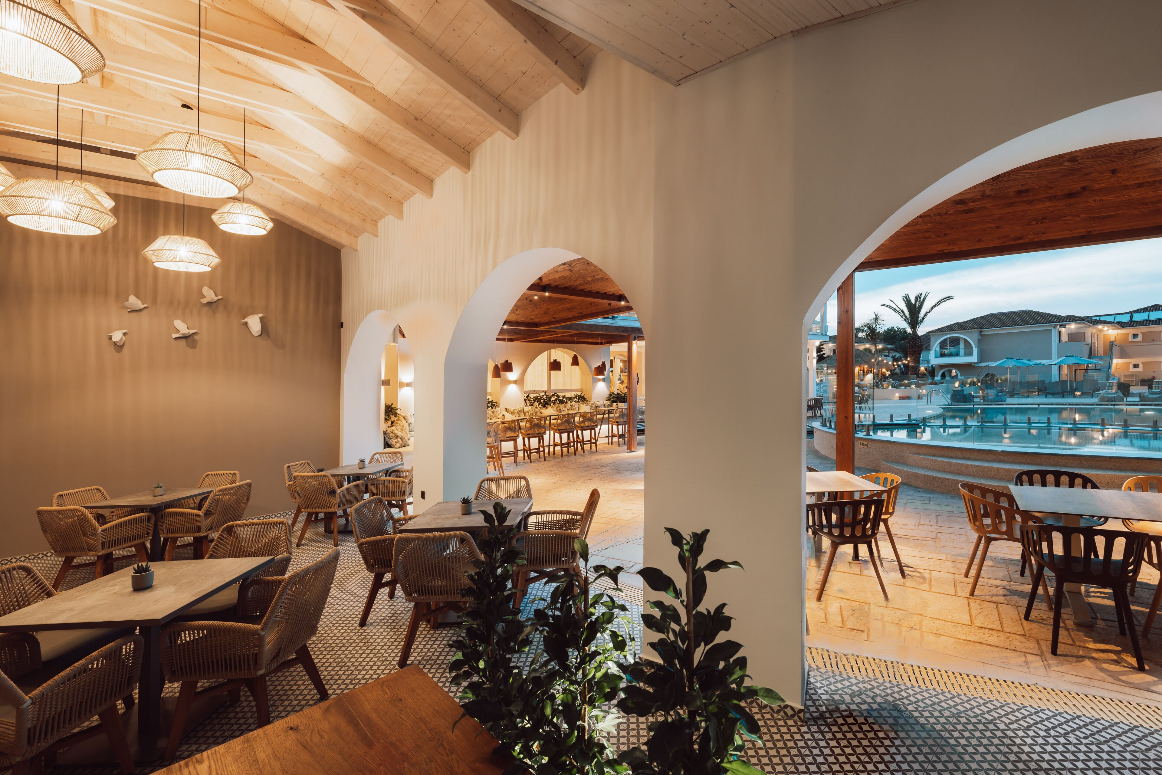A restaurant with wooden ceiling beams, wicker furniture, and a view of a pool and buildings in the background.