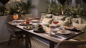 Mesa de comedor servida con platos, vasos y diversos alimentos, rodeada de plantas verdes exuberantes y un diseño interior moderno.