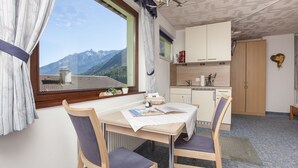 A room with a dining table, chairs, a window with curtains, a kitchen area, and a view of mountains.