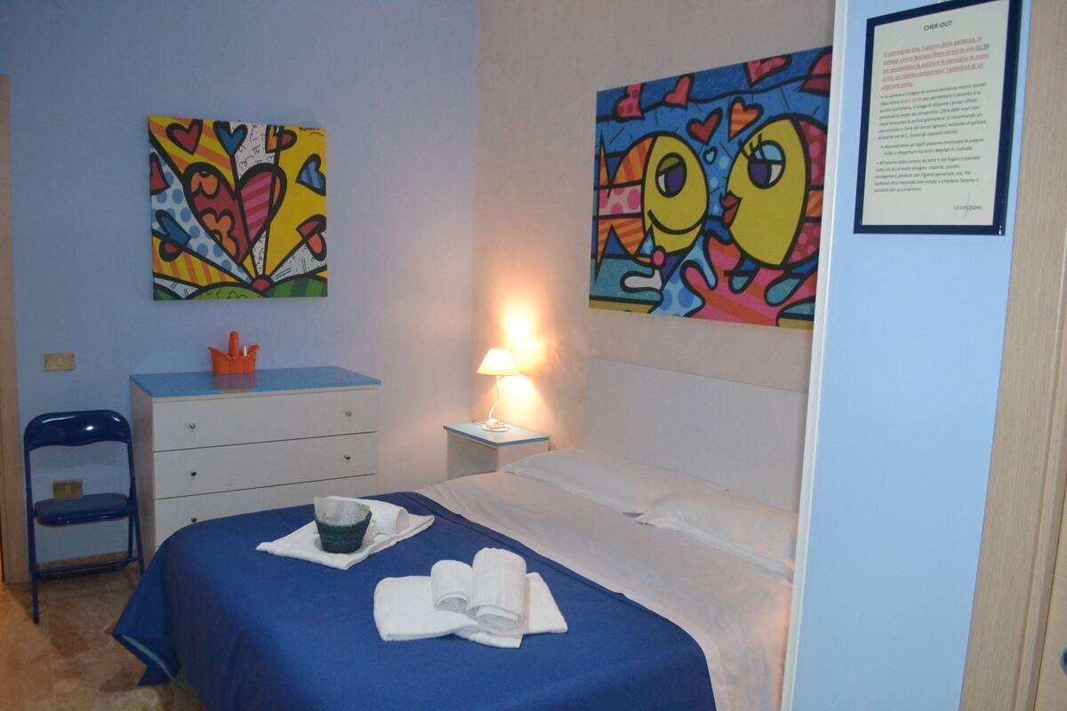 A hotel room with a bed, a desk, a chair, a lamp, and two colorful paintings on the walls.