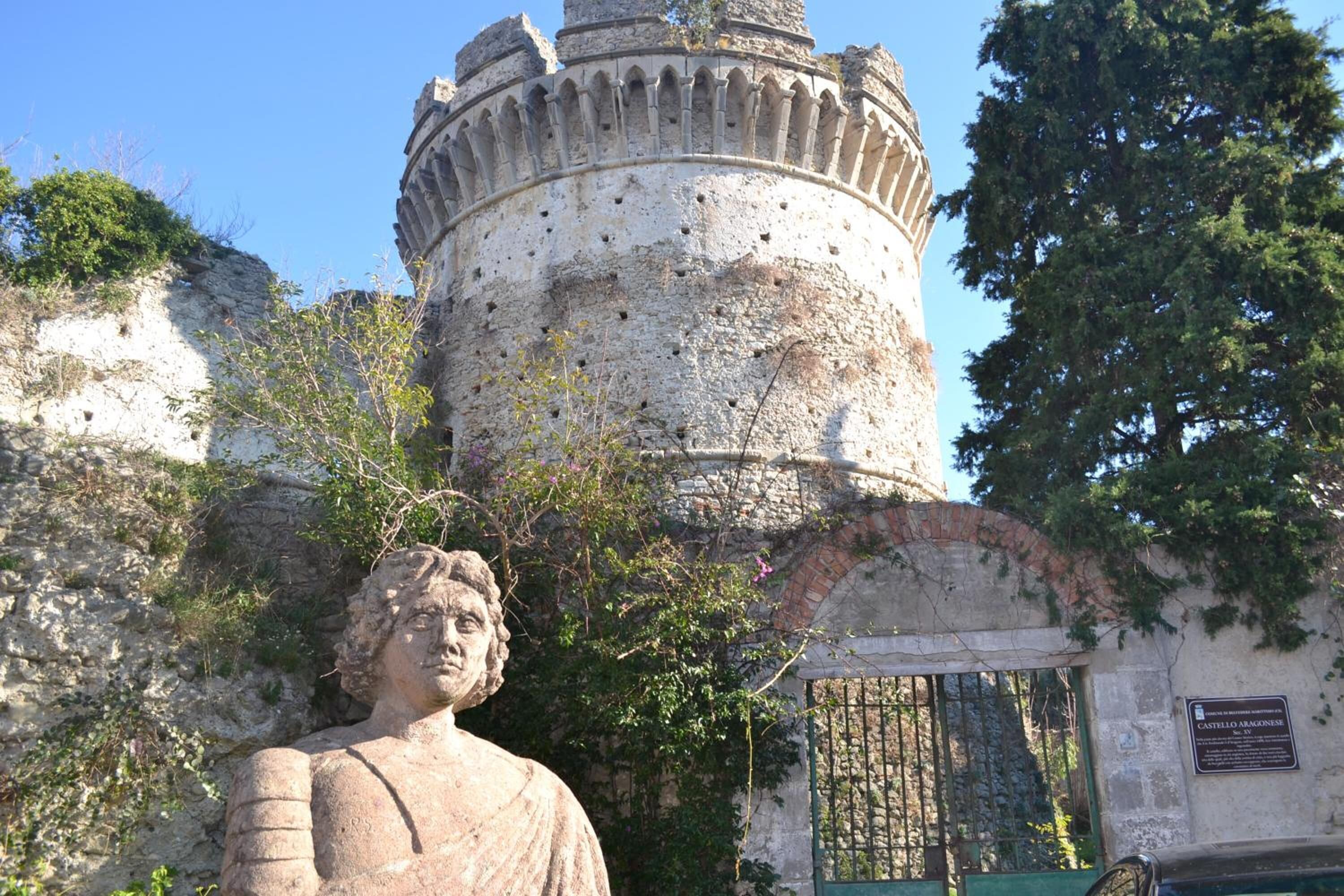 Una estatua de piedra frente a un edificio histórico con una torre redonda y un portón verde.