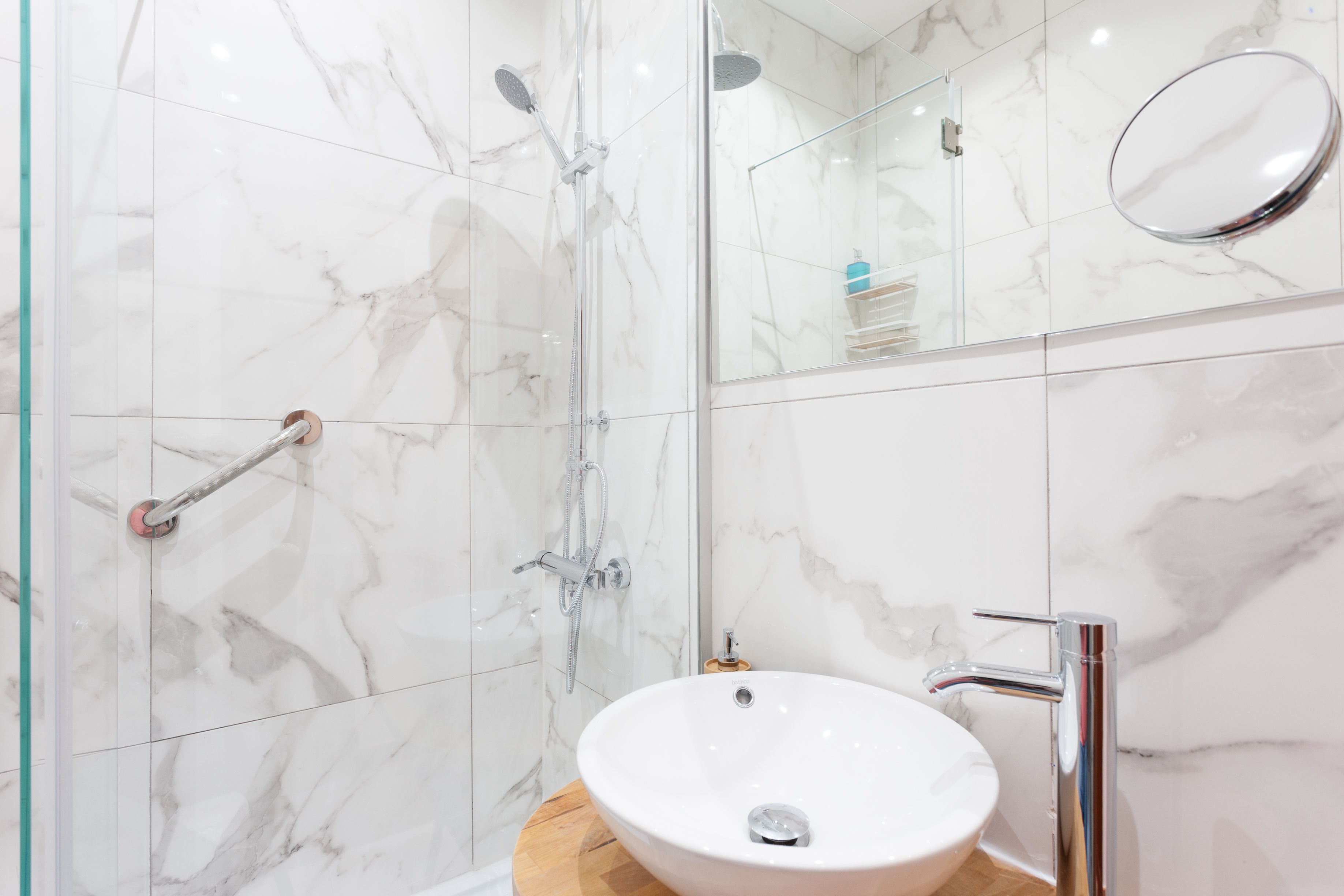 Une salle de bain moderne avec une douche à l’italienne en verre, un lavabo blanc doté d’un plan de toilette en bois et un mur recouvert de carreaux de marbre.