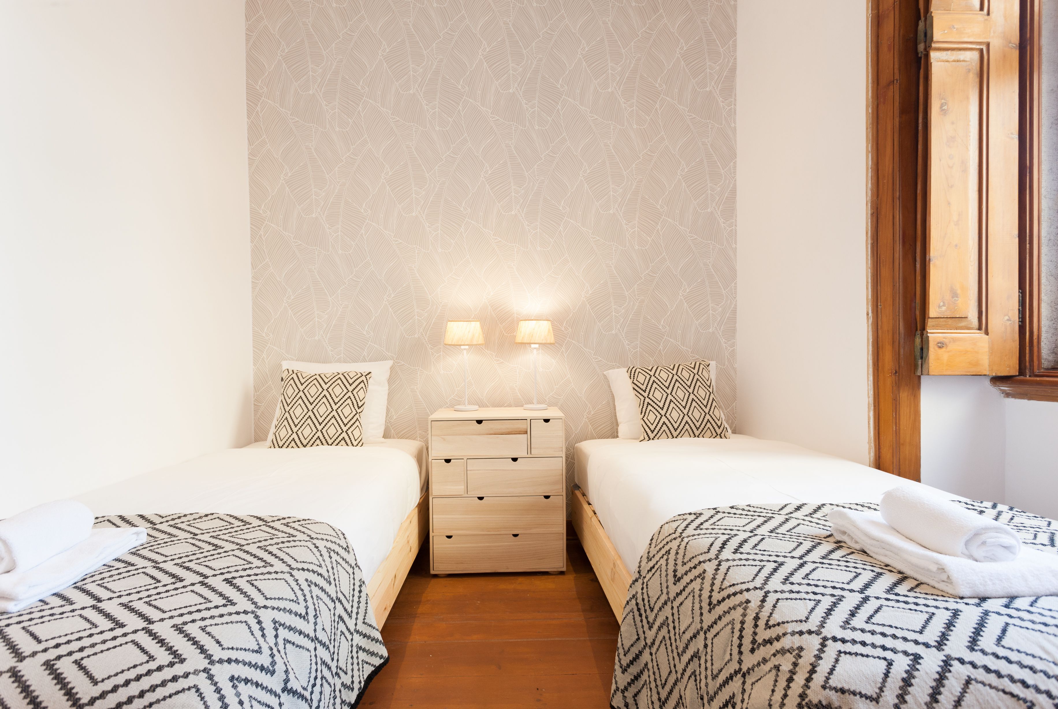 Habitación de hotel con dos camas, una mesita de noche con lámparas, ropa de cama estampada y suelo de madera.