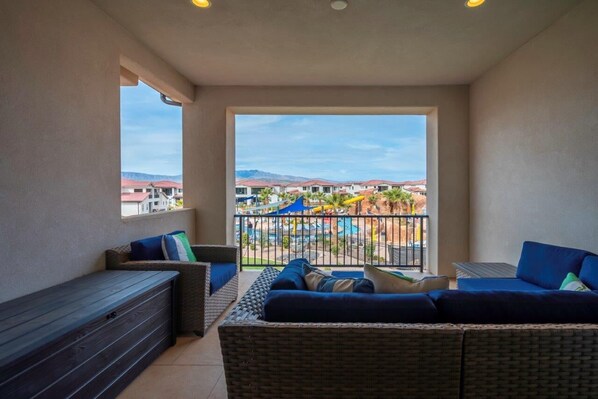 A balcony with wicker furniture, a view of a resort with pools and umbrellas, and a mountain backdrop.