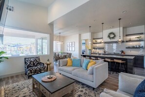 A modern living room with a sofa, coffee table, and a dining area with chairs. There is a kitchen with a bar counter and pendant lights. - Poolside Villa | Air Hockey | Sleeps 18 | Arcadia #3 (Santa Clara)