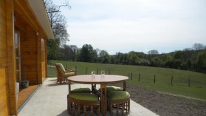Une cabane en bois avec une table ronde et deux chaises en osier dotées de coussins verts, installée sur une terrasse en béton offrant une vue sur un champ verdoyant et des arbres.
