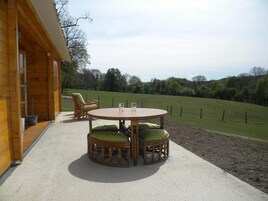 Una cabaña de madera con una mesa redonda y dos sillas de mimbre con cojines verdes, ubicada en un patio de concreto con vista a un campo cubierto de pasto y árboles.