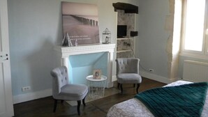 Une pièce avec une cheminée, deux fauteuils, un lit recouvert d’une couverture verte et un tableau accroché au mur.