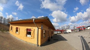 A wooden building with a red roof, a gas station in the background, and a clear blue sky with clouds. - Motel w Lace (Boleslawiec)