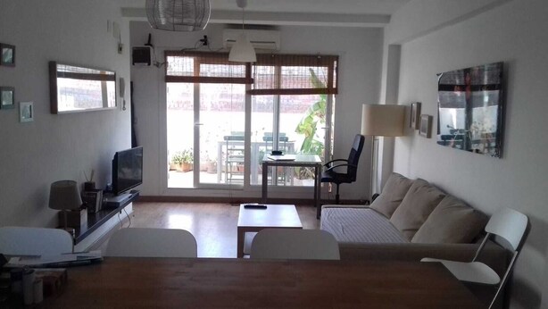 A living room with a sofa, a TV, a table, and a chair. There is a window with blinds and a view of the outside.