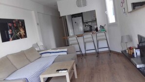 A living room with a sofa, a coffee table, and a TV. There is a kitchen area with a fridge, cabinets, and a window.