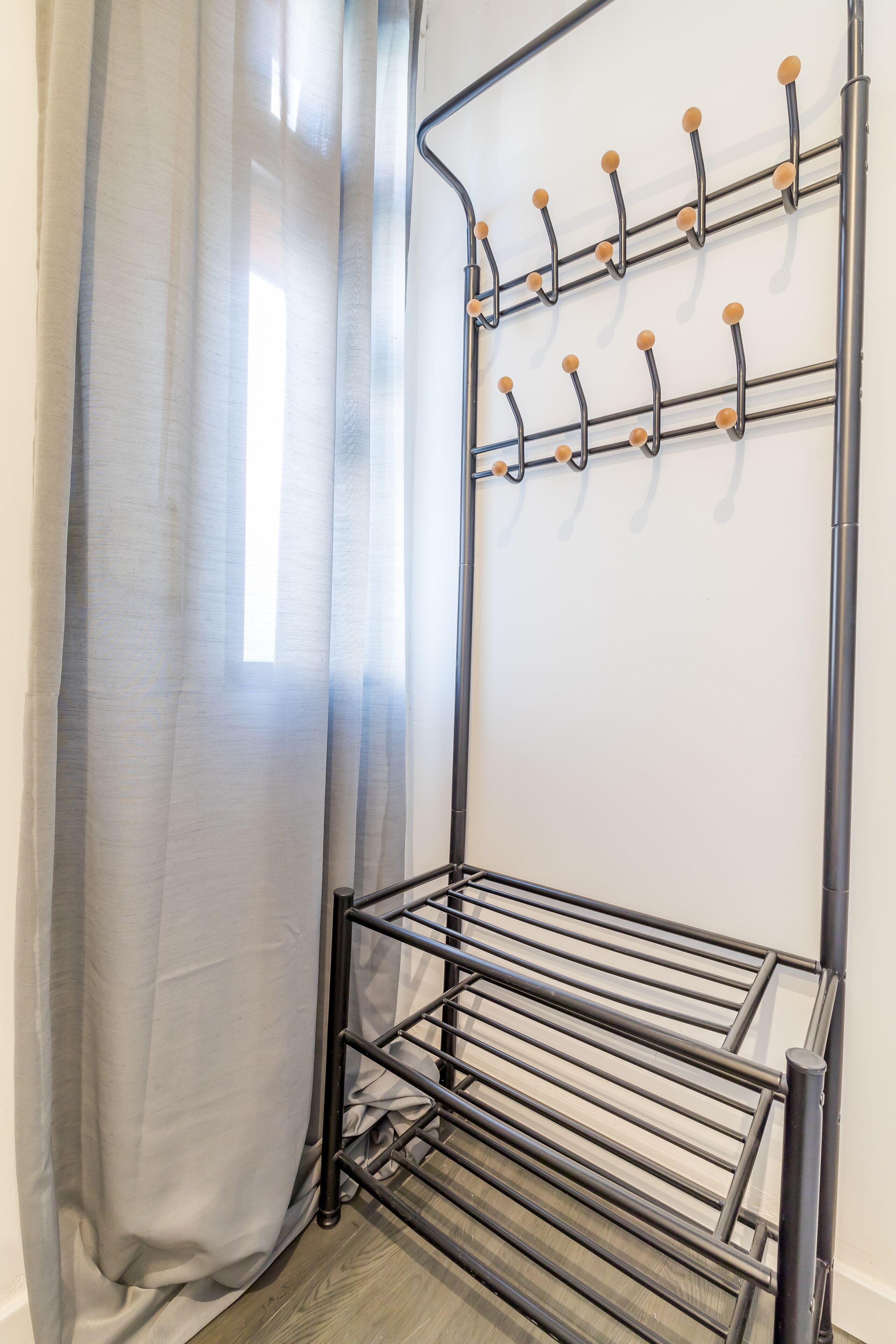 A metal drying rack with multiple hooks and a shelf, positioned against a wall with a window and a curtain.