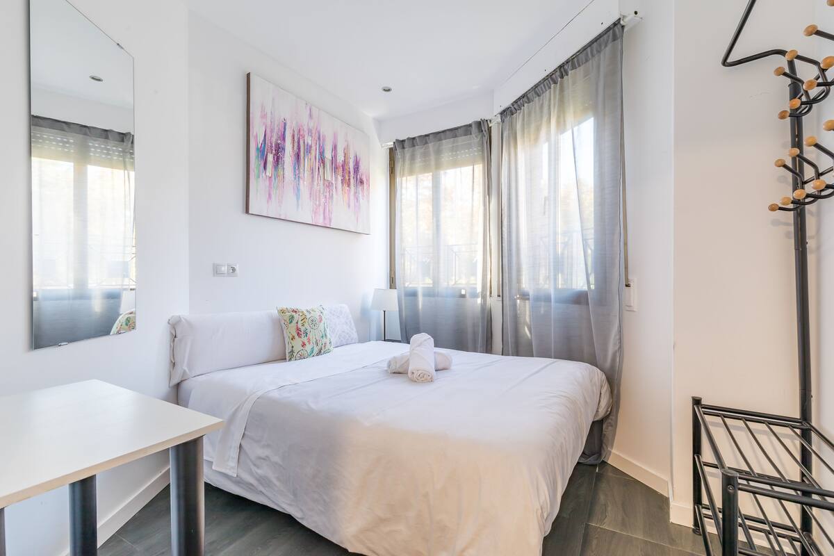Ein Hotelzimmer mit einem Bett, einem Schreibtisch, einem Sessel, einem großen Spiegel, einem Gemälde und einem Fenster mit Vorhängen.