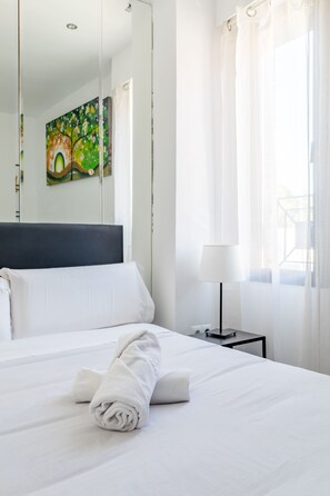 A neatly made bed with white linens and a rolled towel, a bedside table with a lamp, and a painting on the wall. - AYZ Ulises - Auto check-in property (Madrid)
