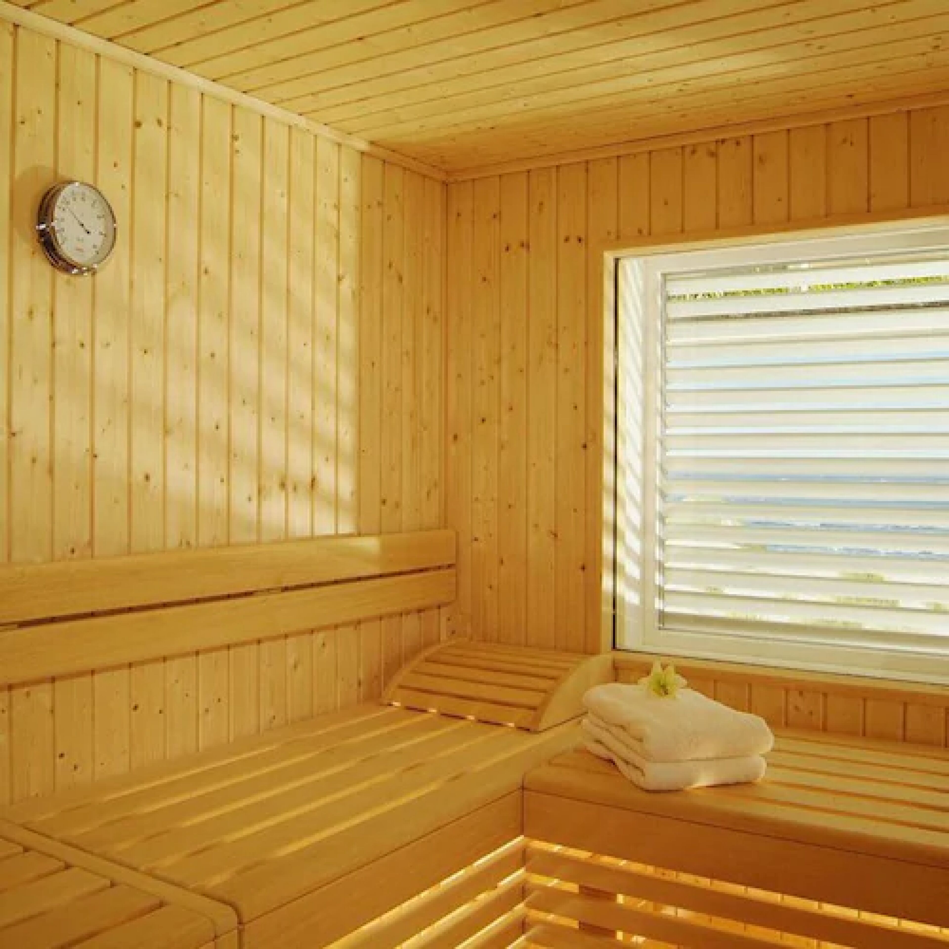 A wooden sauna with benches, a clock, and a window with blinds.