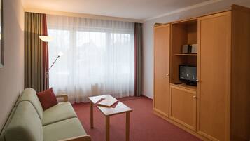 Ein Hotelzimmer mit einem grünen Sofa, einer Holzkabine mit Fernseher, einem kleinen Tisch und einem großen Fenster mit transparenten Vorhängen.