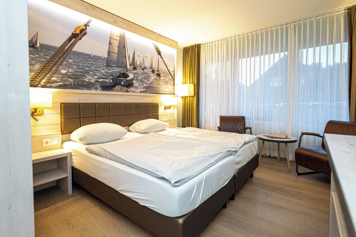 Une chambre d’hôtel avec un grand lit, un bureau, une chaise et un tableau représentant un pont et des voiliers accroché au mur.