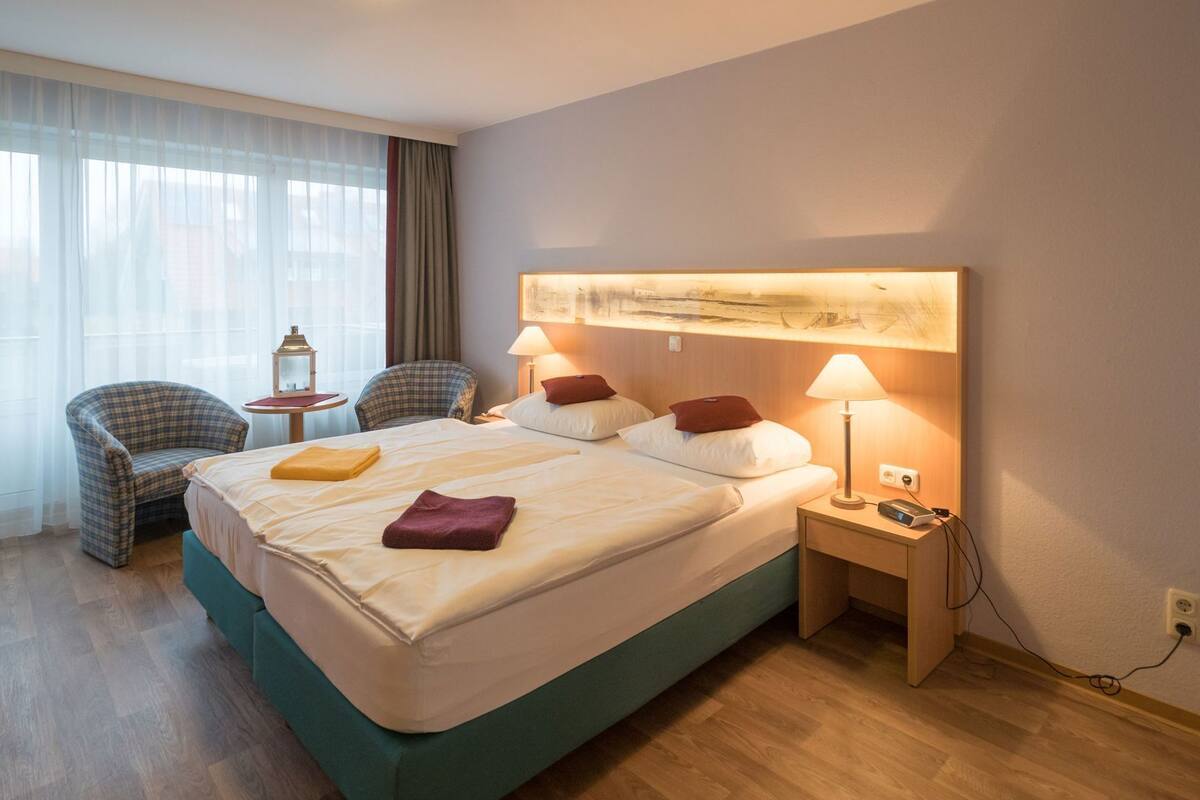 Une chambre d’hôtel avec un lit, deux chaises, une petite table, une lampe et un tableau au mur.