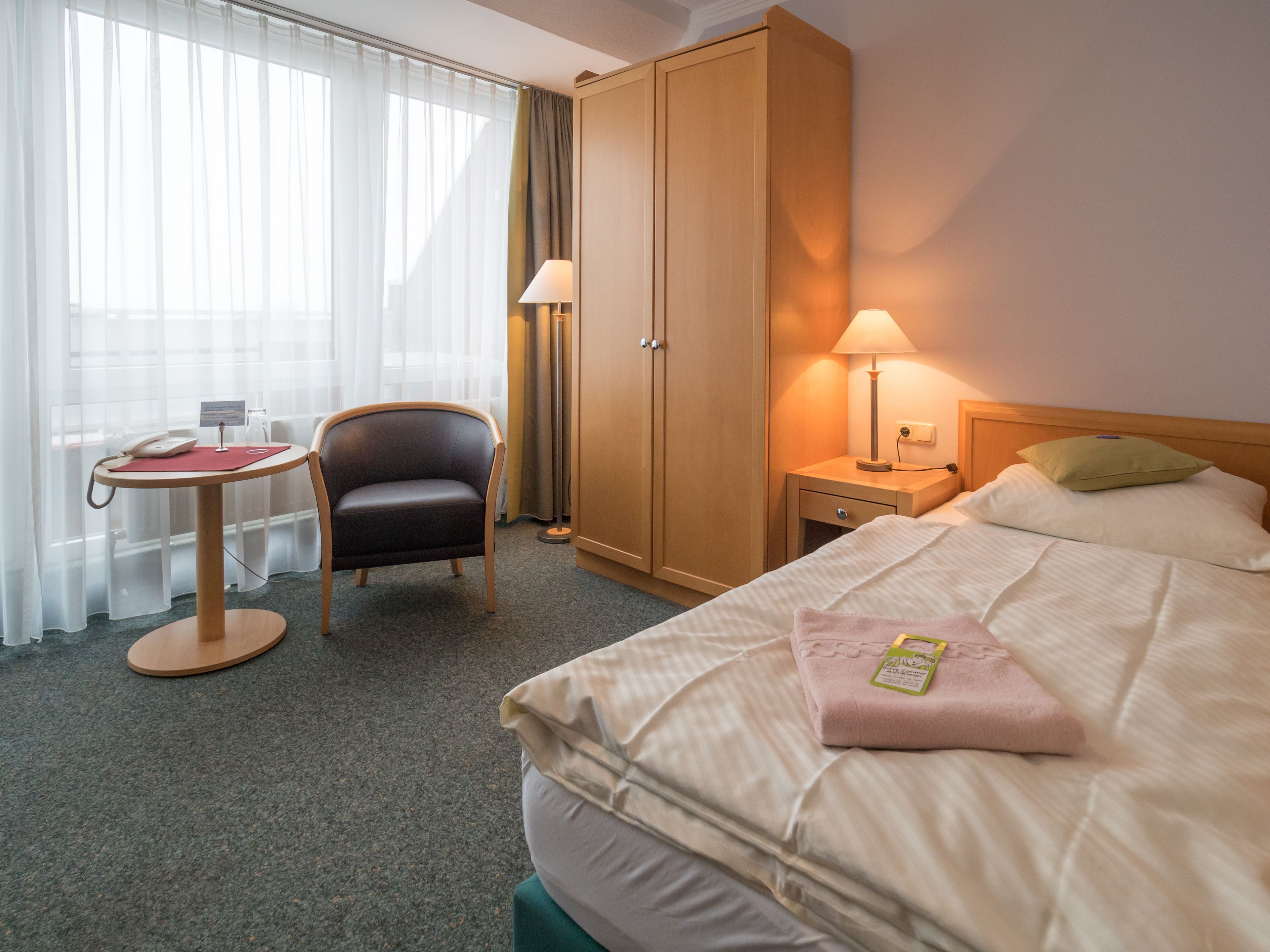 A hotel room with a bed, a desk with a chair, a wardrobe, and a bedside table with a lamp.