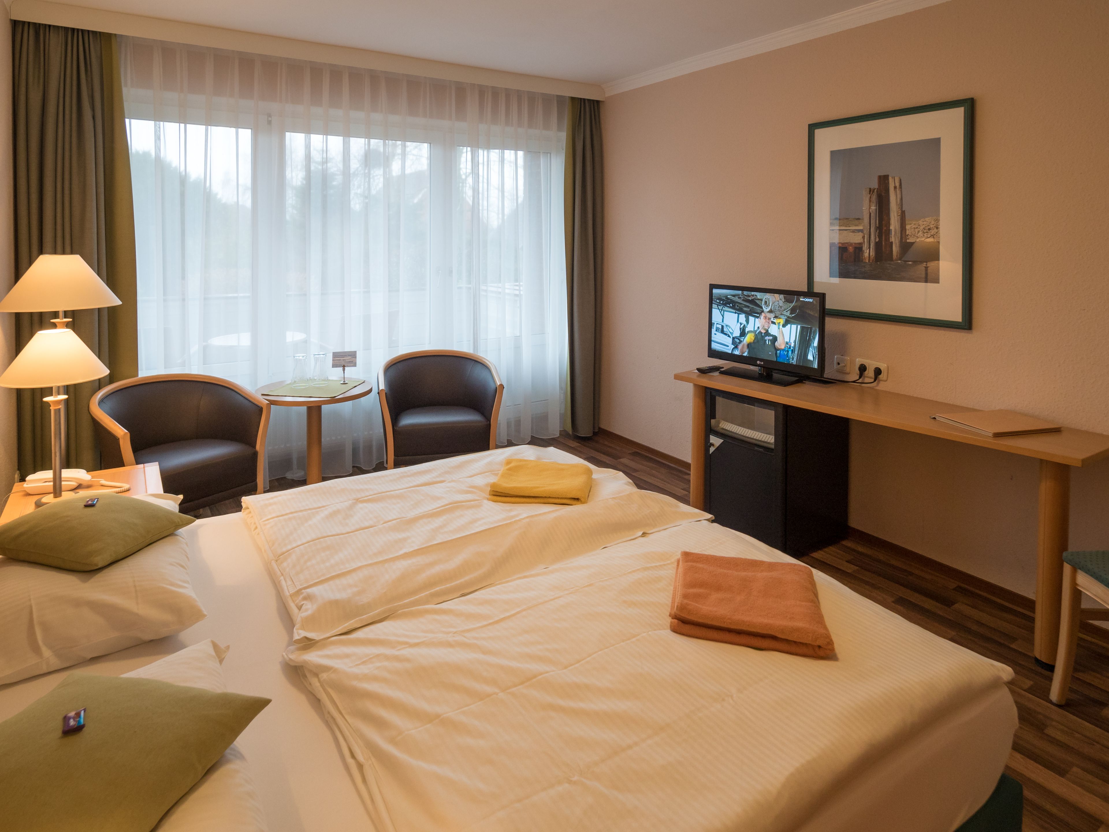 A hotel room with a bed, a desk with a television, two chairs, a lamp, and a framed picture on the wall.