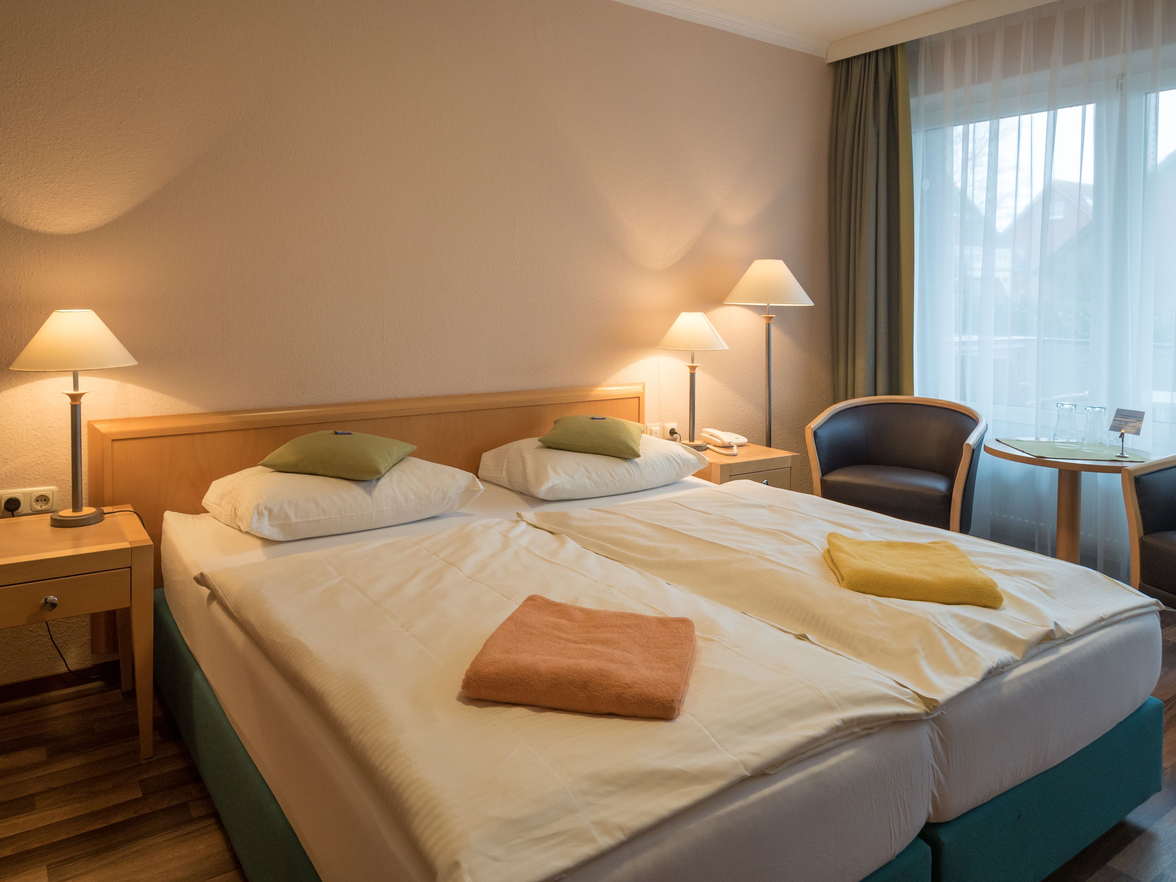 A hotel room with two beds, a chair, a small table, and two lamps.