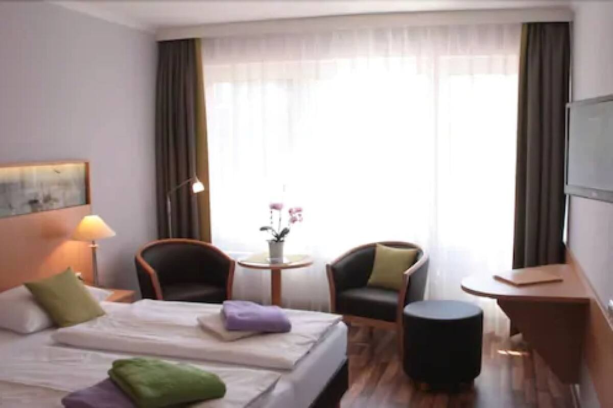 Une chambre d’hôtel avec un lit, deux chaises, une petite table avec un vase, une lampe et un téléviseur.