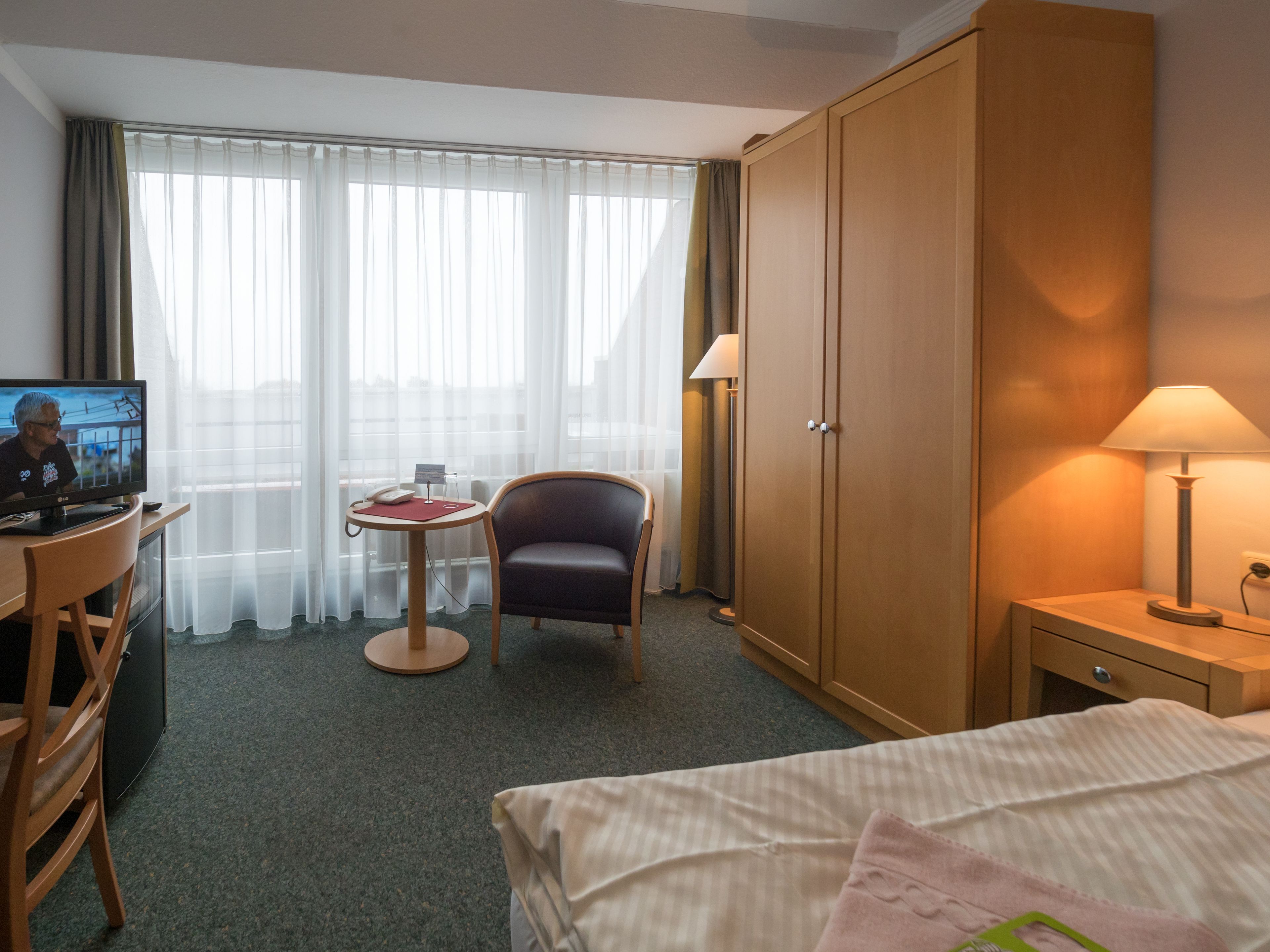 Et hotelværelse med en seng, et skrivebord med tv, en stol, et klædeskab og et natbord med lampe.