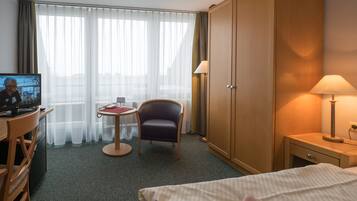 Et hotelværelse med en seng, et skrivebord med tv, en stol, et klædeskab og et natbord med lampe.