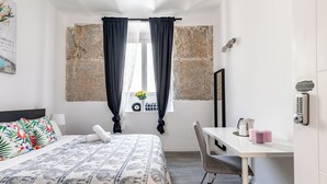 Ein Hotelzimmer mit einem Bett, einem Schreibtisch, einem Sessel, einem Fenster mit Vorhängen, einer Steinwand und einer Uhr.
