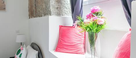 Quarto de hotel bem arrumado com uma cama, uma janela com cortinas, um travesseiro rosa, um vaso com flores e uma luminária.