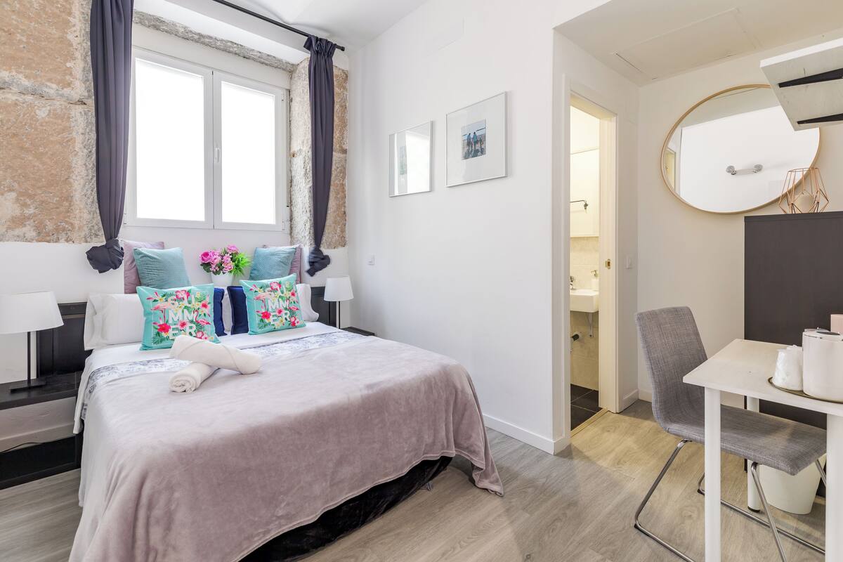 Una habitación de hotel moderna con cama, escritorio, silla y un baño visible a través de una puerta abierta.