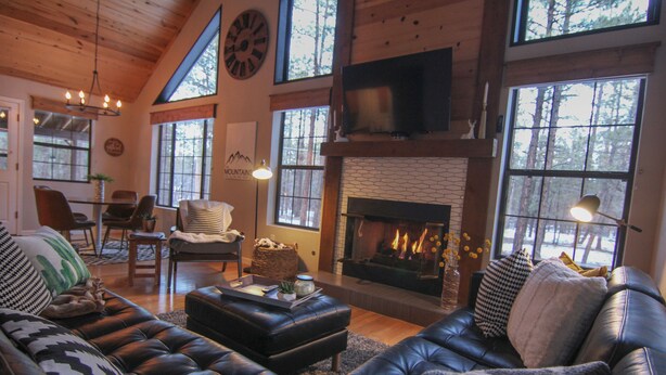A cozy living room with leather sofas, a fireplace, and large windows offering a view of the outdoors. - Luxury Cabin Retreat with Hot Tub on Spacious Wooded Lot (Pinetop)