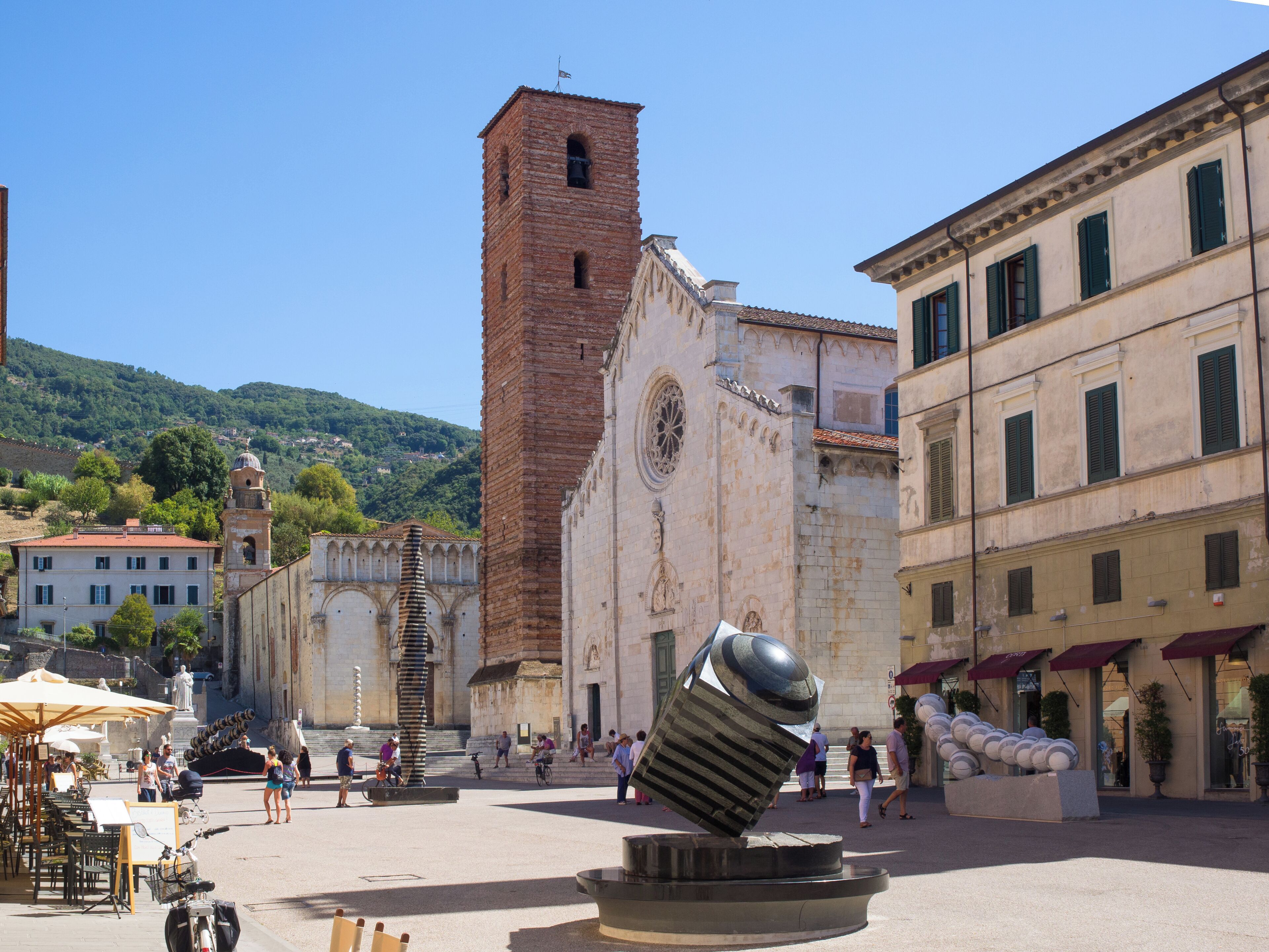 A square with a large, tilted sculpture in the center, surrounded by historic buildings and a church with a bell tower.