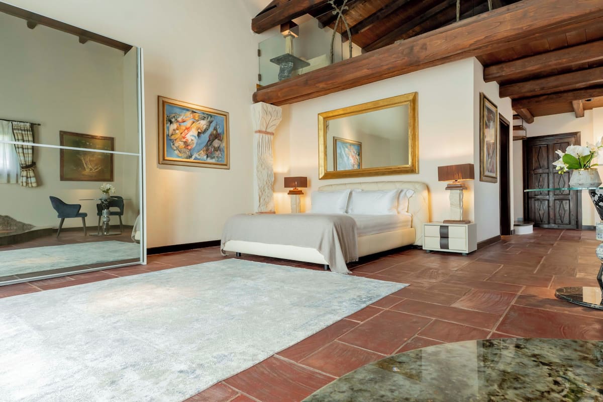 A bedroom with a bed, bedside lamps, a large mirror, and a view of a dining area.