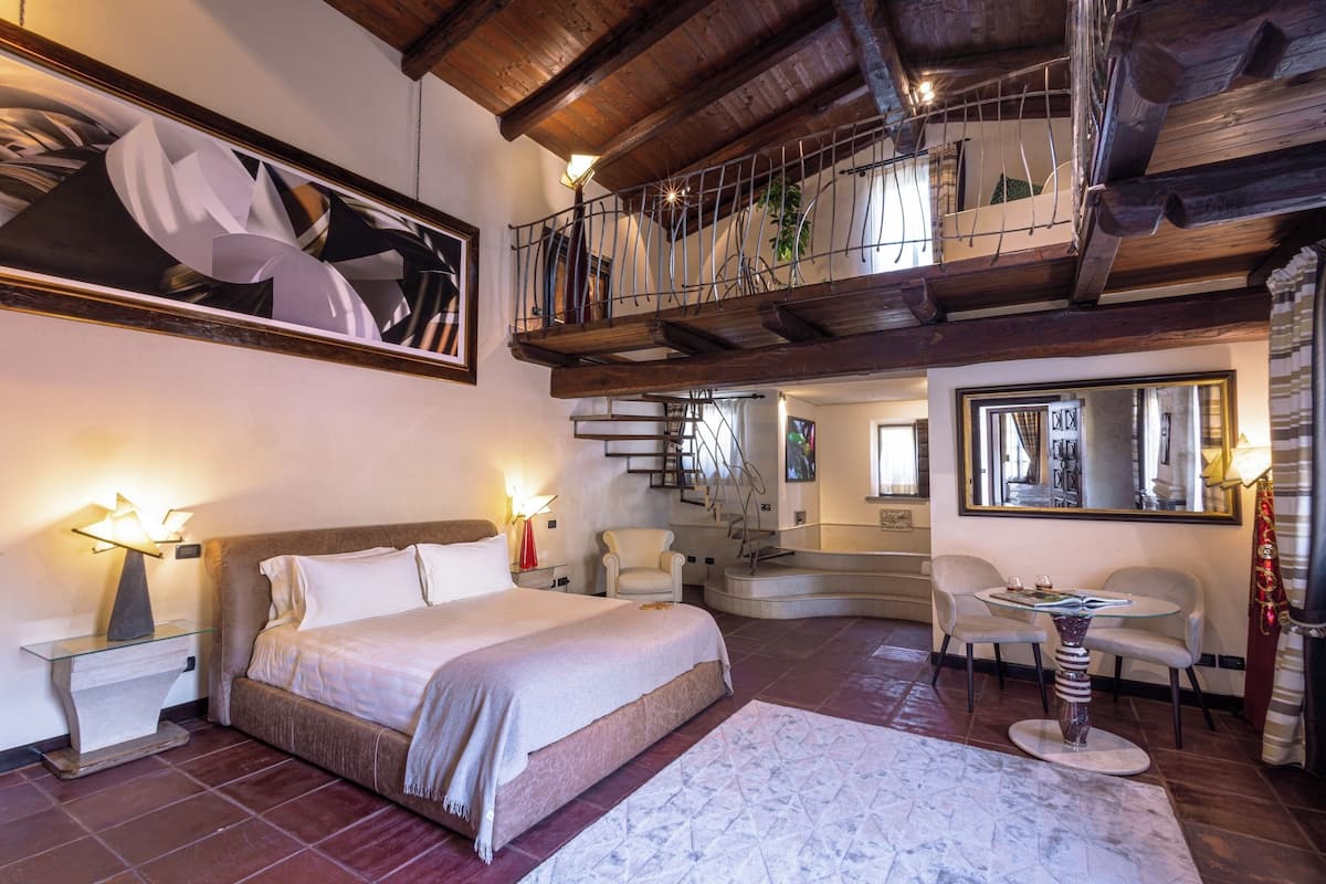 A bedroom with a large bed, a glass table, a chair, a staircase, and a balcony.
