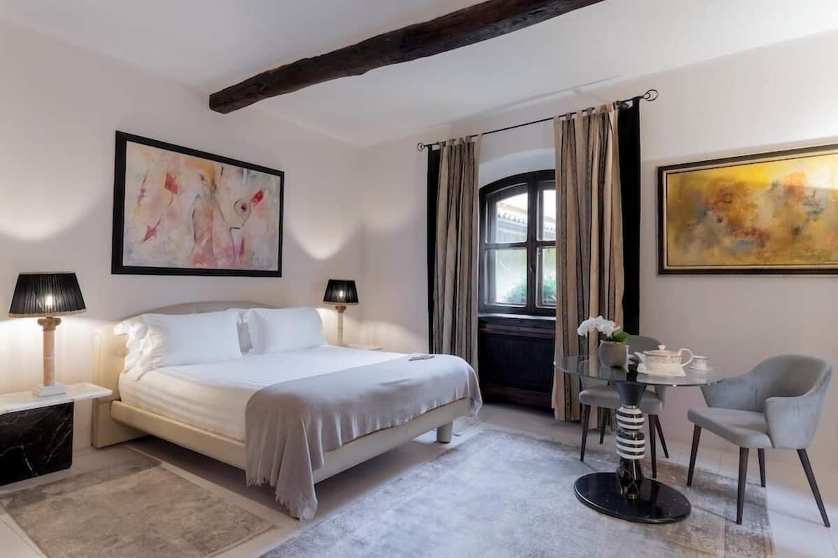 A bedroom with a bed, bedside lamps, a glass table with chairs, and artwork on the walls.