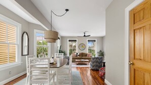 A spacious living room with a wooden floor, a dining area with white wicker furniture, and a large window with blinds. - Your Rooftop Haven | Sunset Suite Serenity (Charleston)