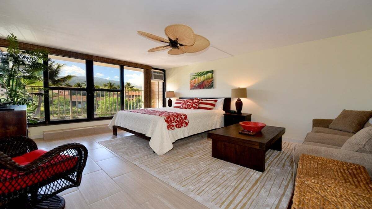 A spacious bedroom with a large bed, a ceiling fan, a balcony with a view of palm trees, and a sitting area with a sofa and a coffee table.