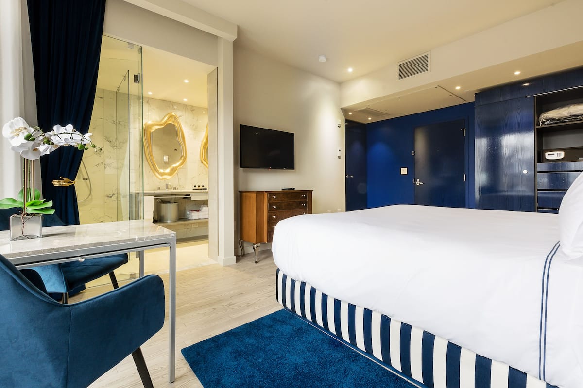 A modern hotel room with a large bed, a desk with a chair, a TV, and a bathroom with a glass shower.
