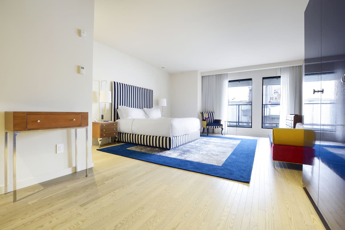 A modern hotel room with a large bed, a desk, and a chair. There are windows with curtains and a view of the outside.