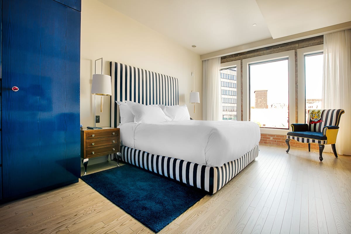 A modern hotel room with a large bed, a blue wardrobe, a nightstand with a lamp, and a view of the city through large windows.
