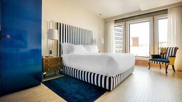 A modern hotel room with a large bed, a blue wardrobe, a nightstand with a lamp, and a view of the city through large windows.