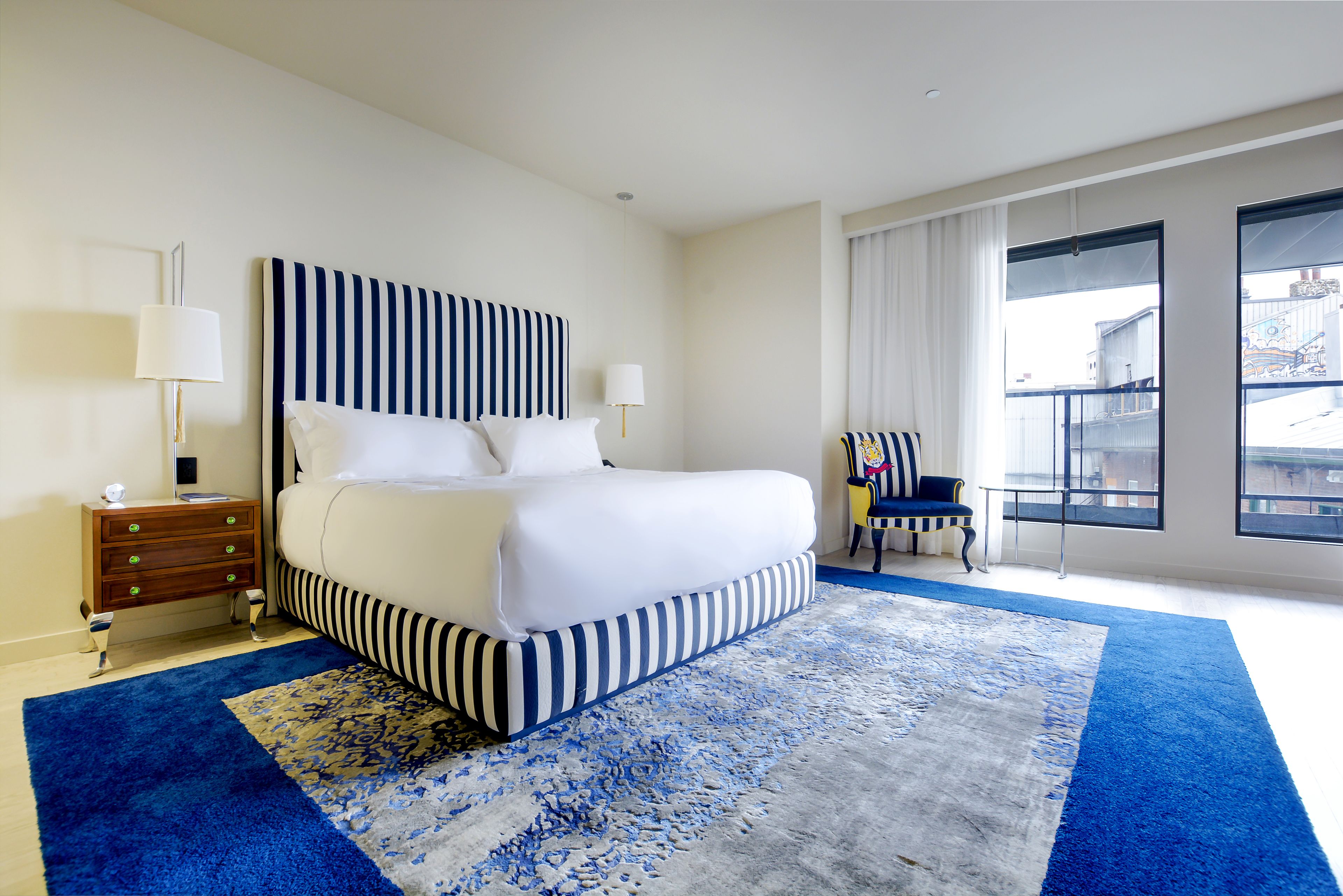 A hotel room with a large bed, a nightstand, a chair, and a view of the city through large windows.